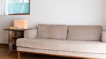 A beige sofa with a woven texture, a small round table with a basket and a folded orange towel, and a window with a view of trees.