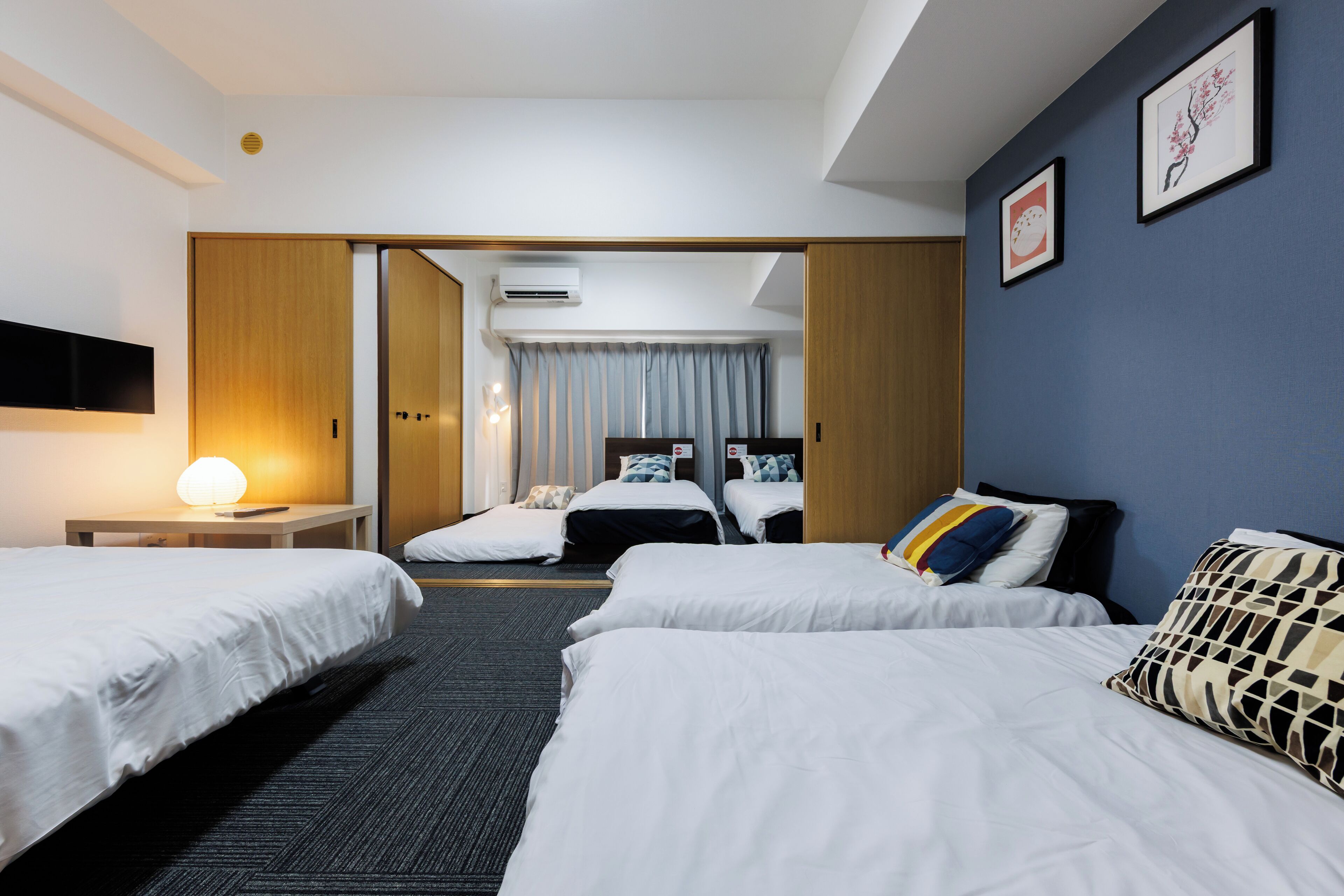 Una habitación de hotel con tres camas individuales, un televisor de pantalla plana, un escritorio con lámpara y un ventanal con cortinas.