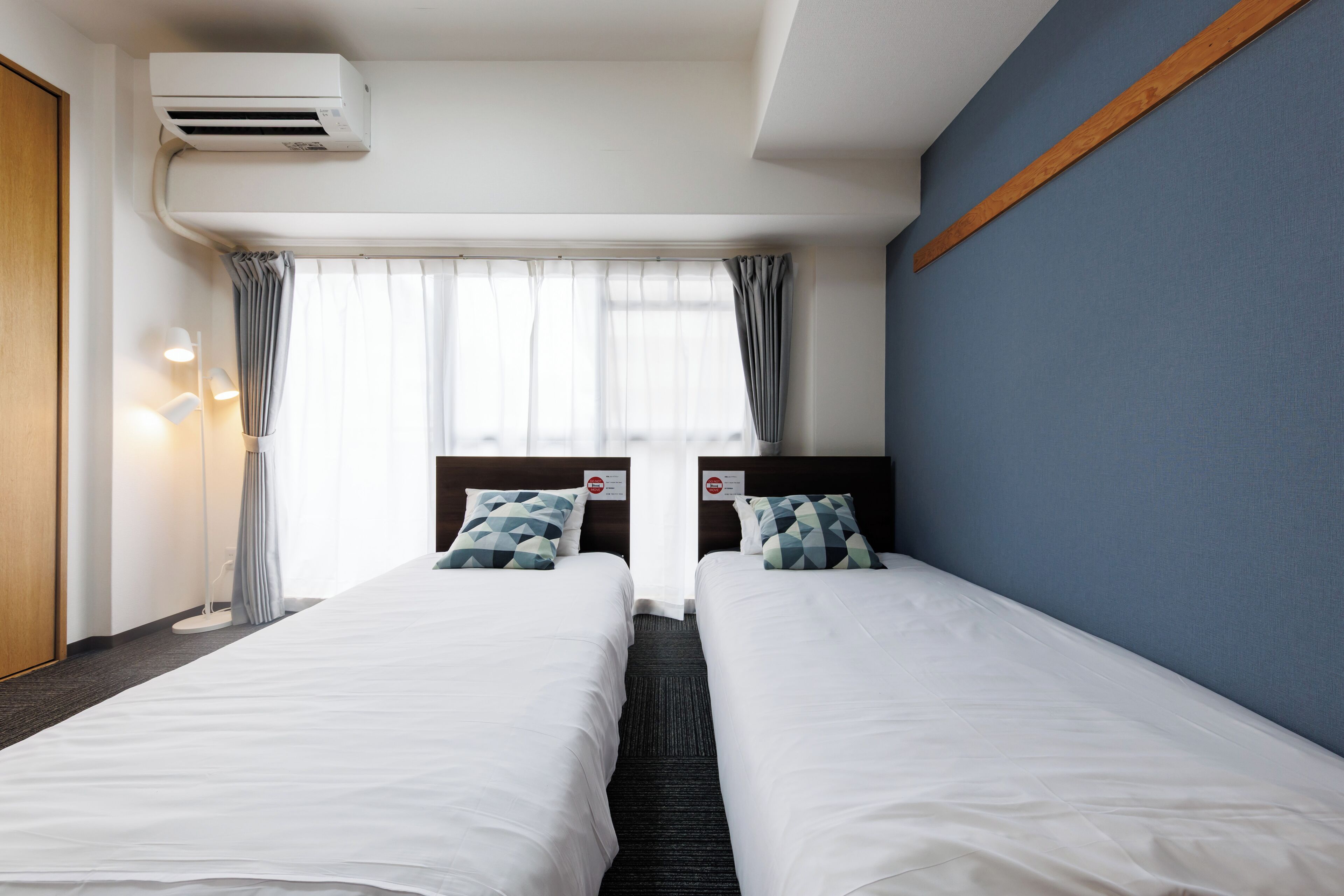 A hotel room with two beds, a window with curtains, a wall-mounted air conditioner, and a bedside lamp.