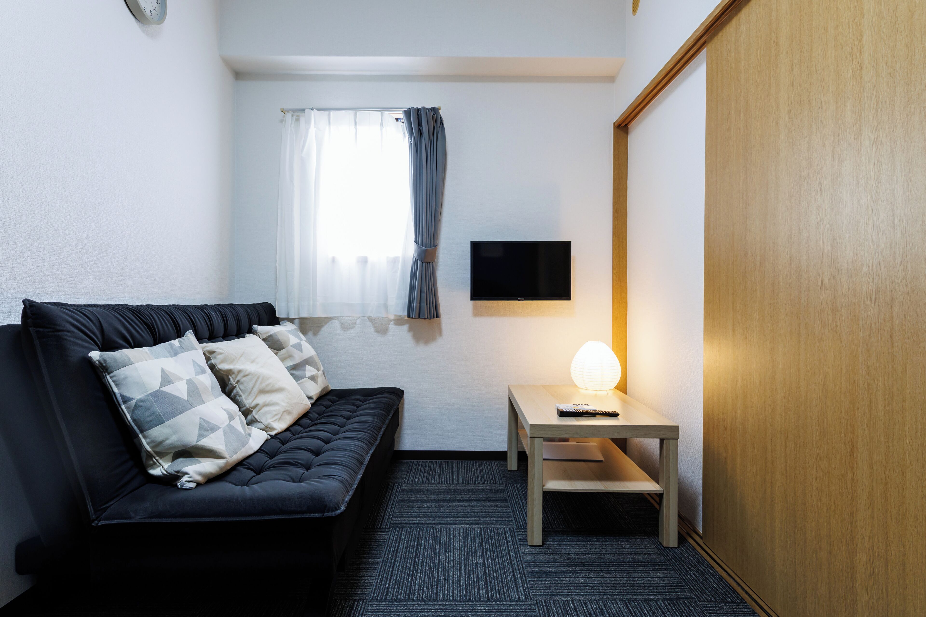 Een moderne hotelkamer met een zwarte bank, een klein houten tafeltje met een lamp, een flatscreen televisie aan de muur en een raam met dunne gordijnen.