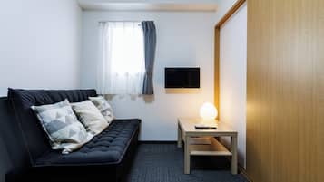 Een moderne hotelkamer met een zwarte bank, een klein houten tafeltje met een lamp, een flatscreen televisie aan de muur en een raam met dunne gordijnen.