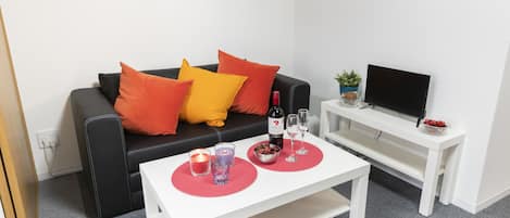 A modern living room with a black leather sofa, red and yellow cushions, a white coffee table with candles, a wine bottle, and a TV on a white stand.