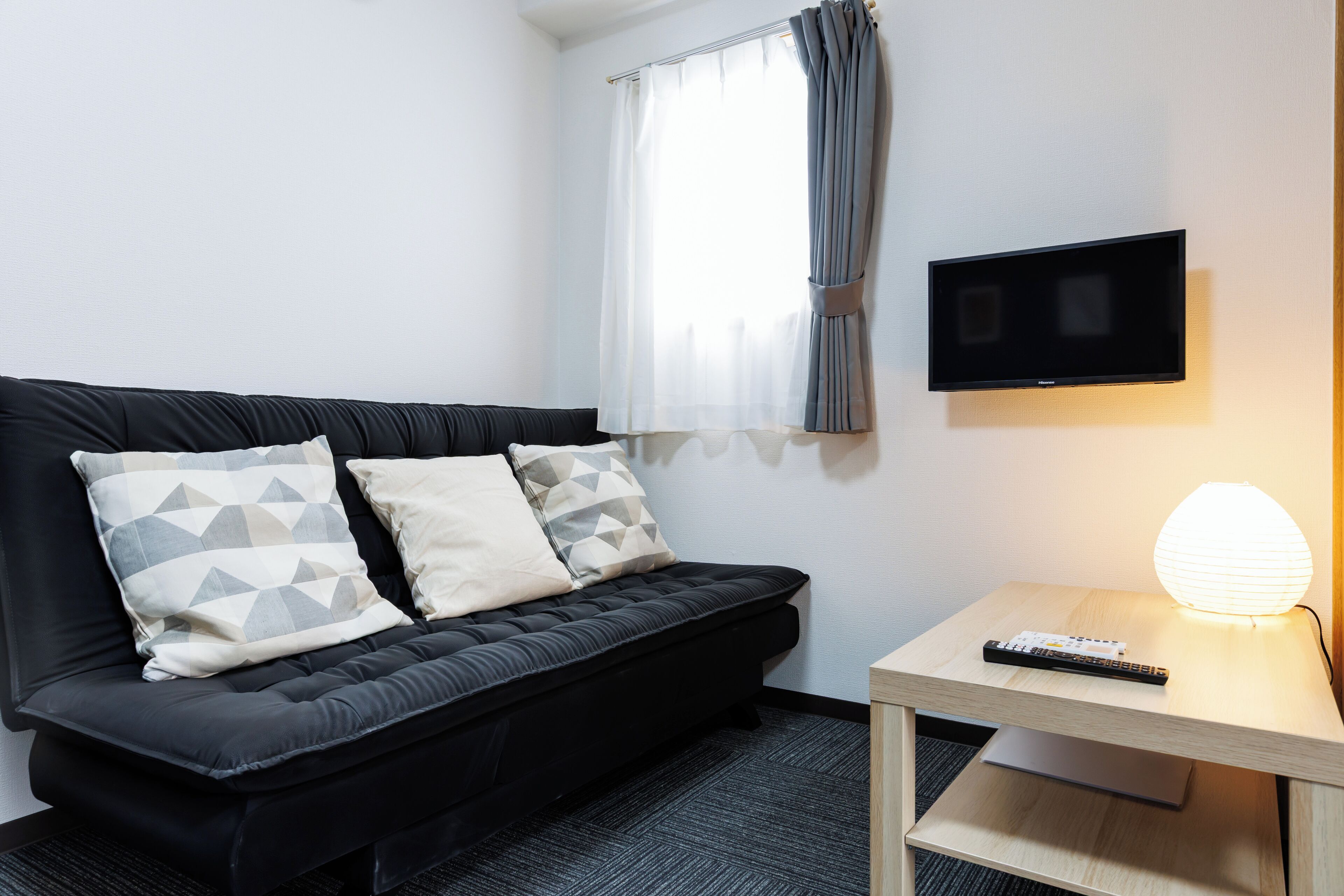 Une pièce moderne avec un canapé noir, un téléviseur à écran plat fixé au mur, une petite table d’appoint en bois avec une télécommande et une lampe.