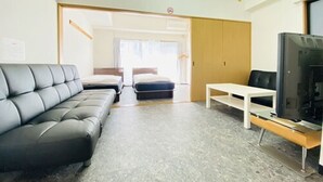 Una habitación de hotel moderna con un sofá de cuero gris, dos camas individuales, una mesita blanca y un televisor.