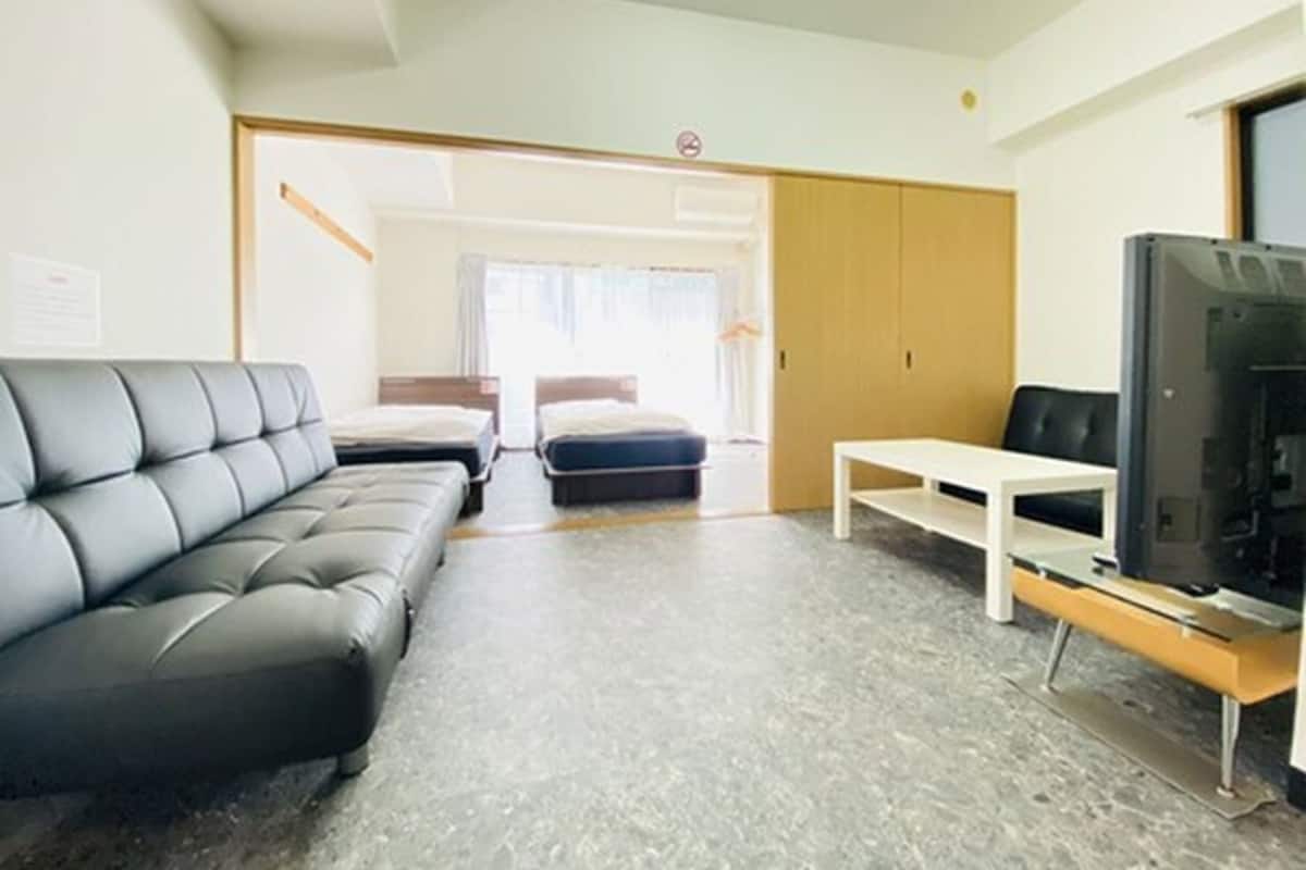 A modern hotel room with a grey leather sofa, two single beds, a small white table, and a TV.