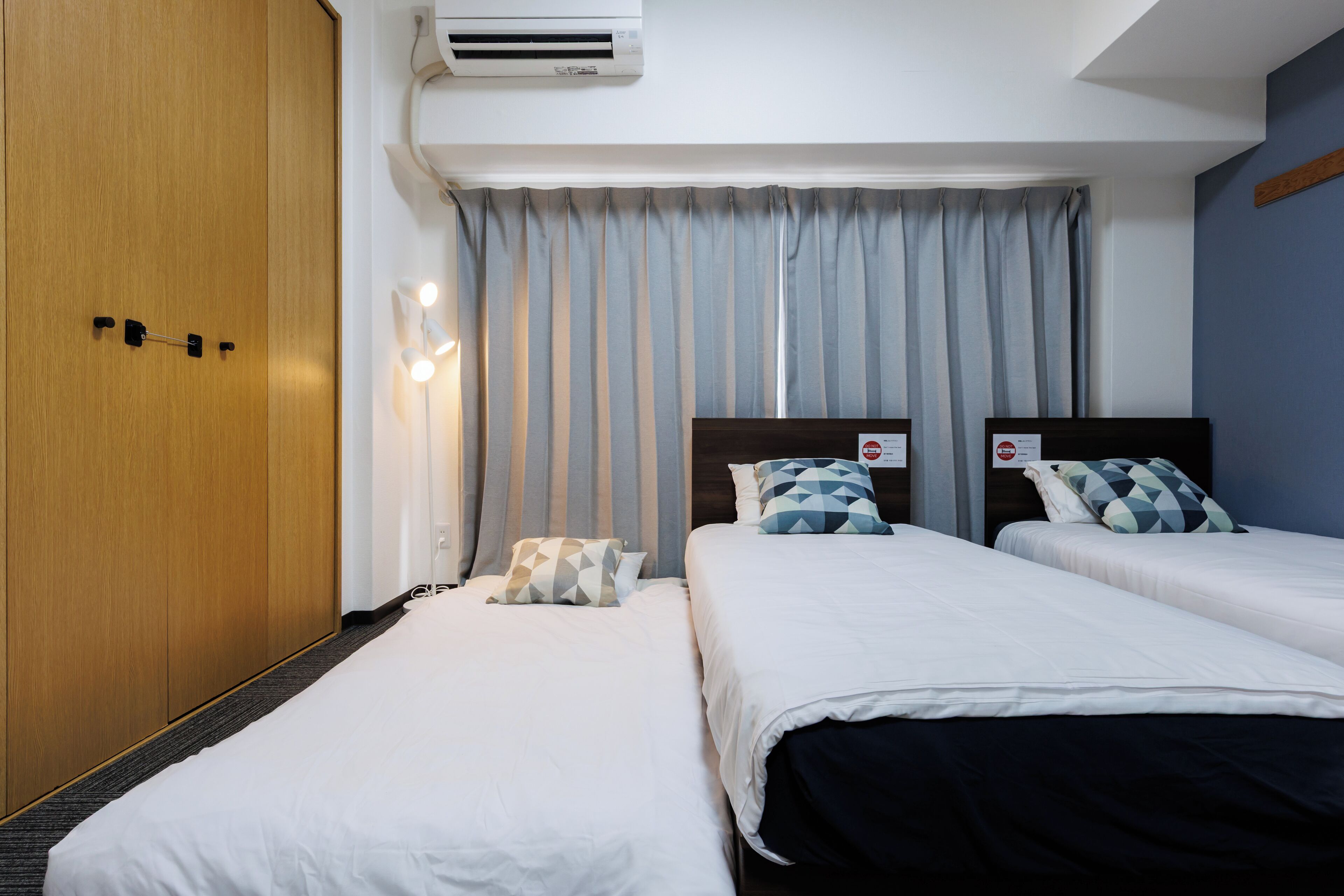 A hotel room with two beds, a wardrobe, a window with curtains, and an air conditioning unit.