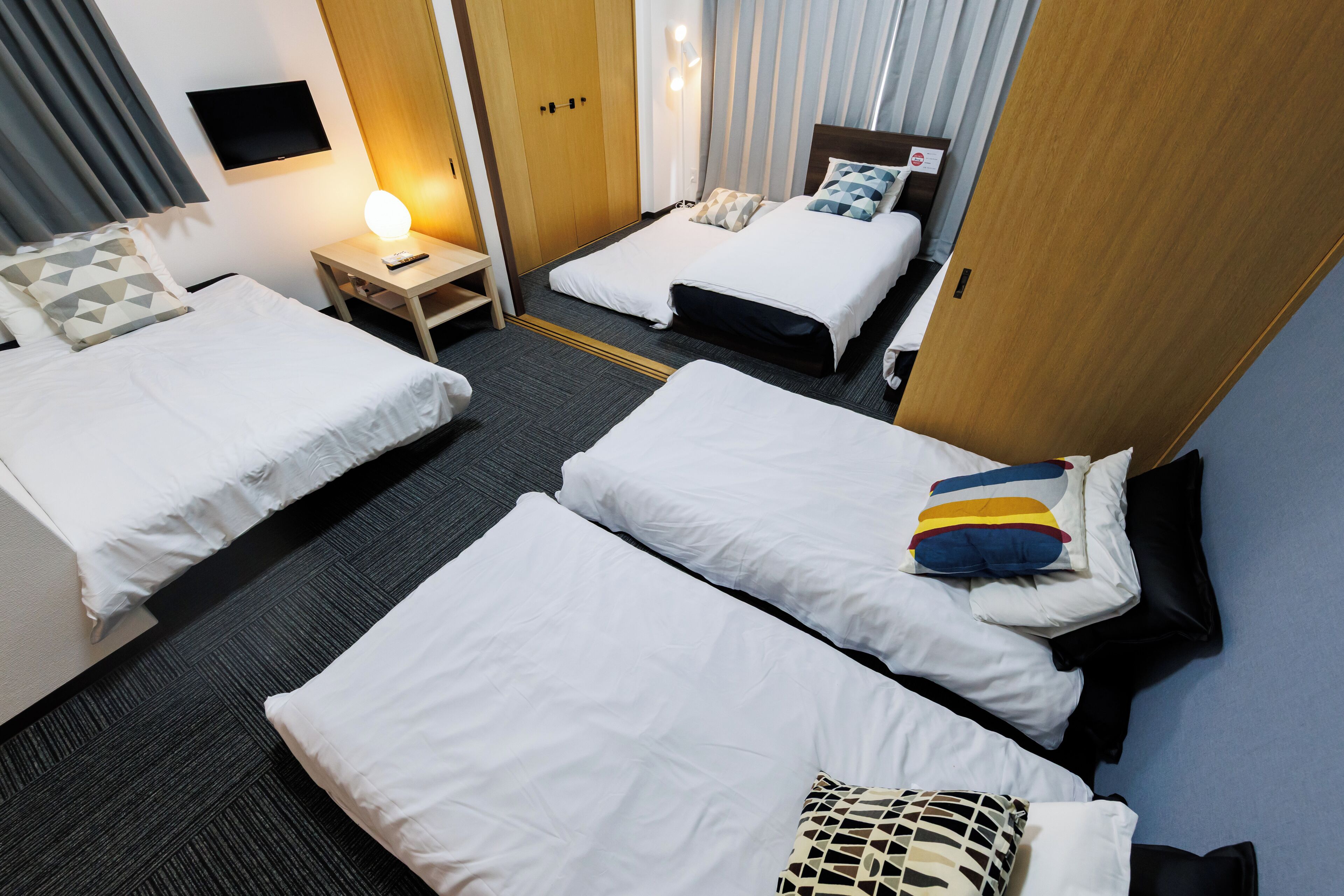 A hotel room with two single beds, a desk with a lamp, a TV on the wall, and a wooden wardrobe.