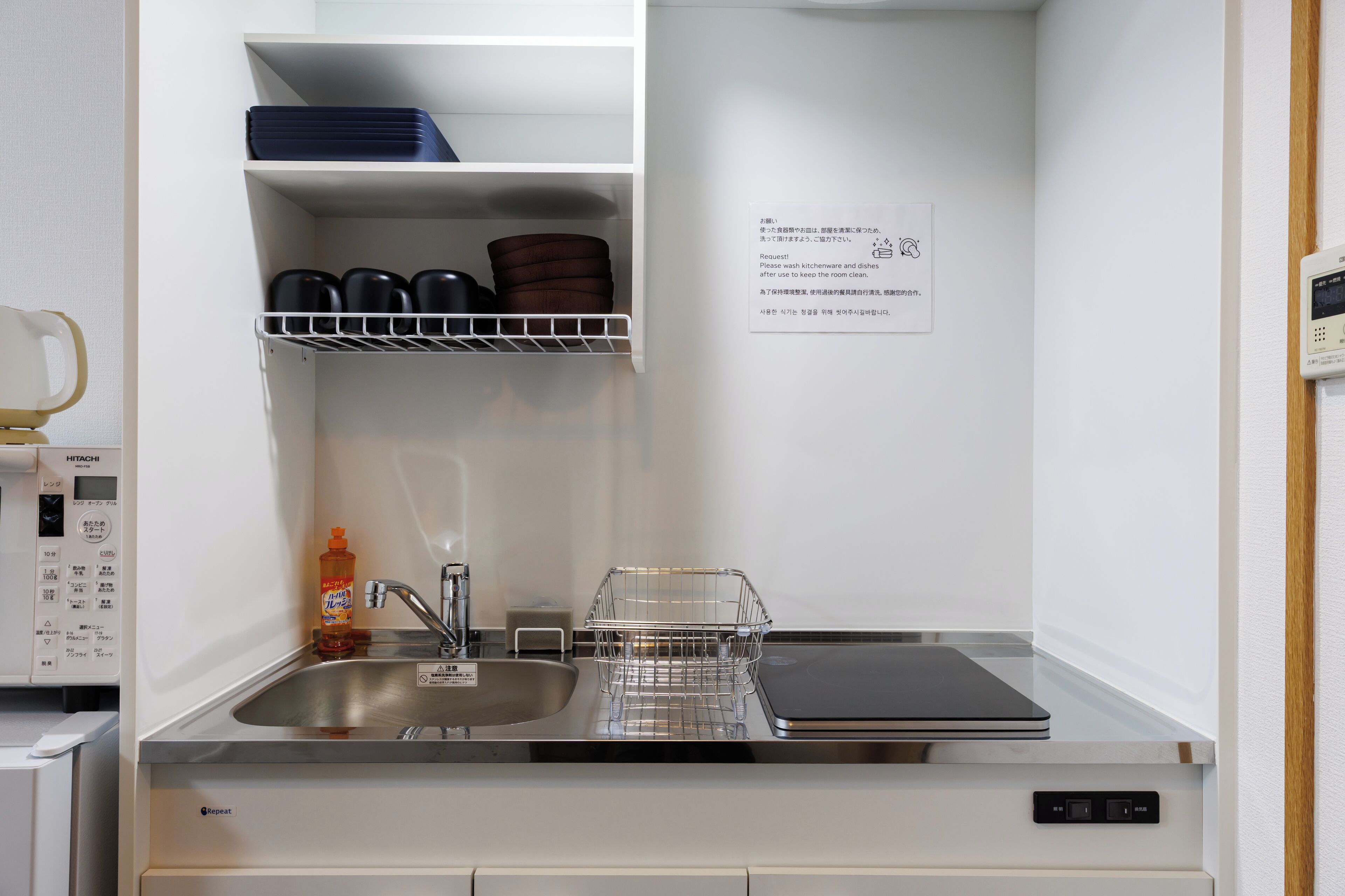 A modern kitchen with a stainless steel sink, a dishwasher, and a built-in oven. There is a shelf above the sink holding kitchenware and a note on the wall.