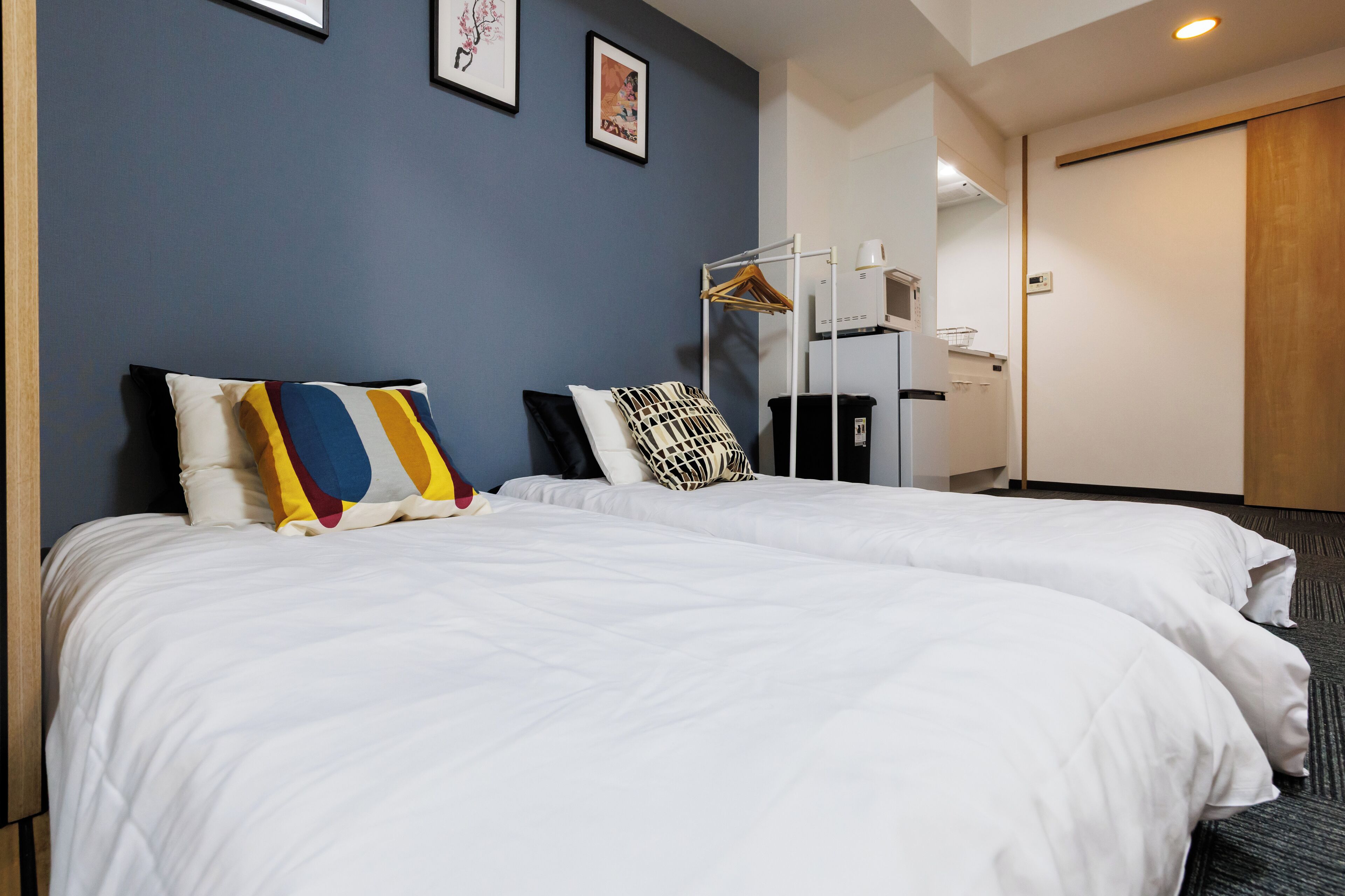 A hotel room with two beds, a wardrobe, a wardrobe with hanging clothes, and a trash bin.