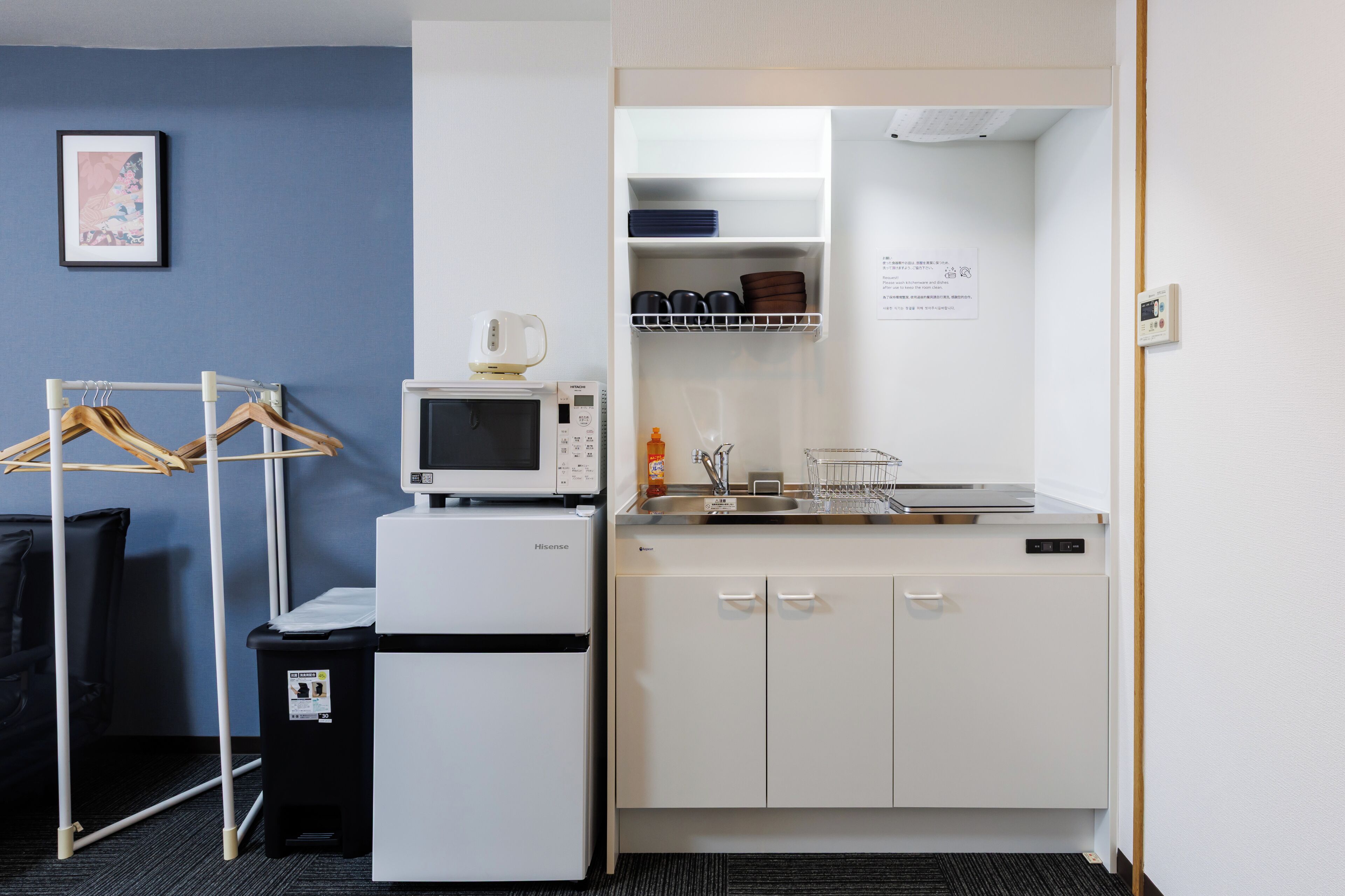 A compact kitchenette with a microwave, small refrigerator, and a sink. There is a drying rack and a trash bin.