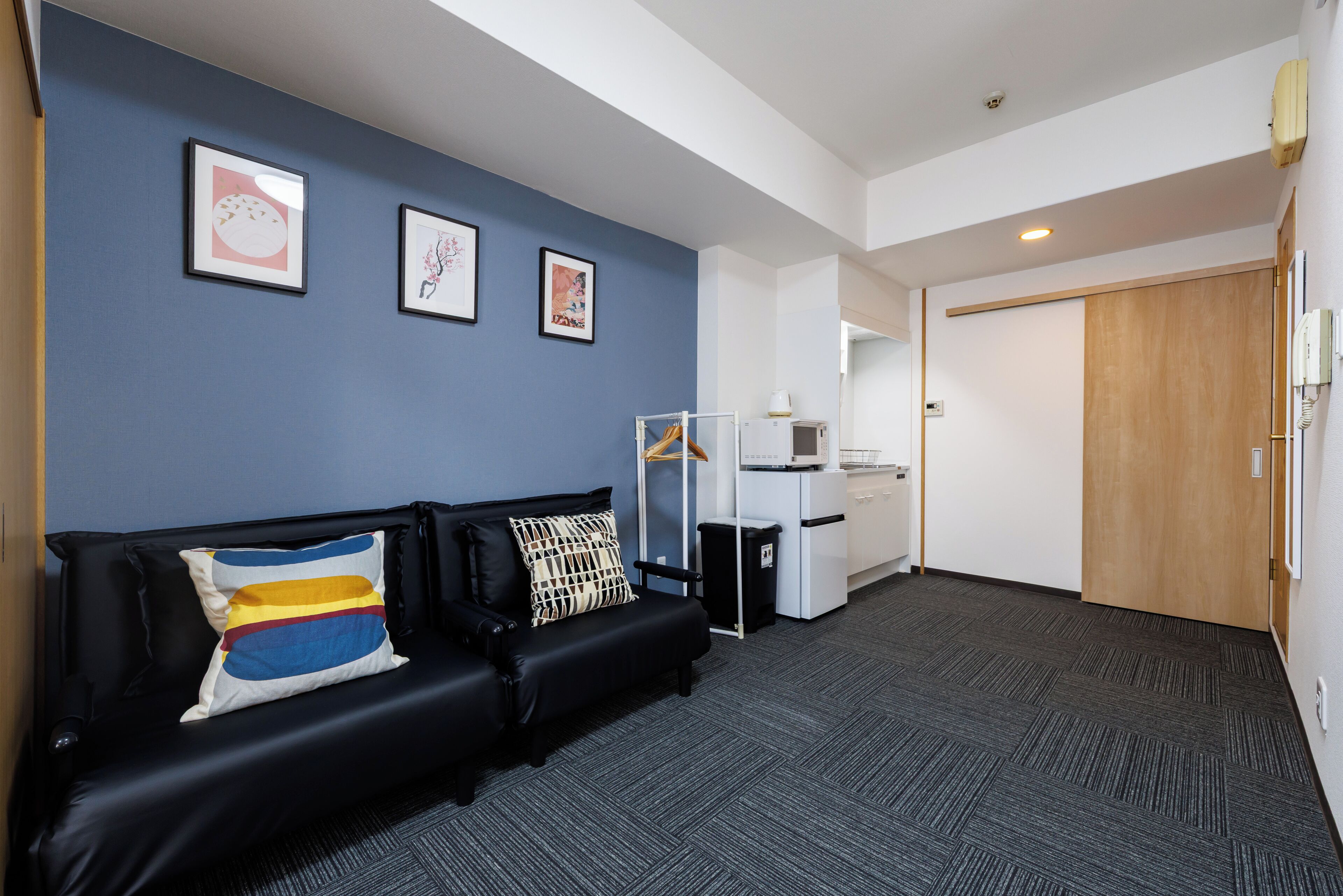 A modern hotel room with a black leather sofa, a small kitchenette, and a wooden door.