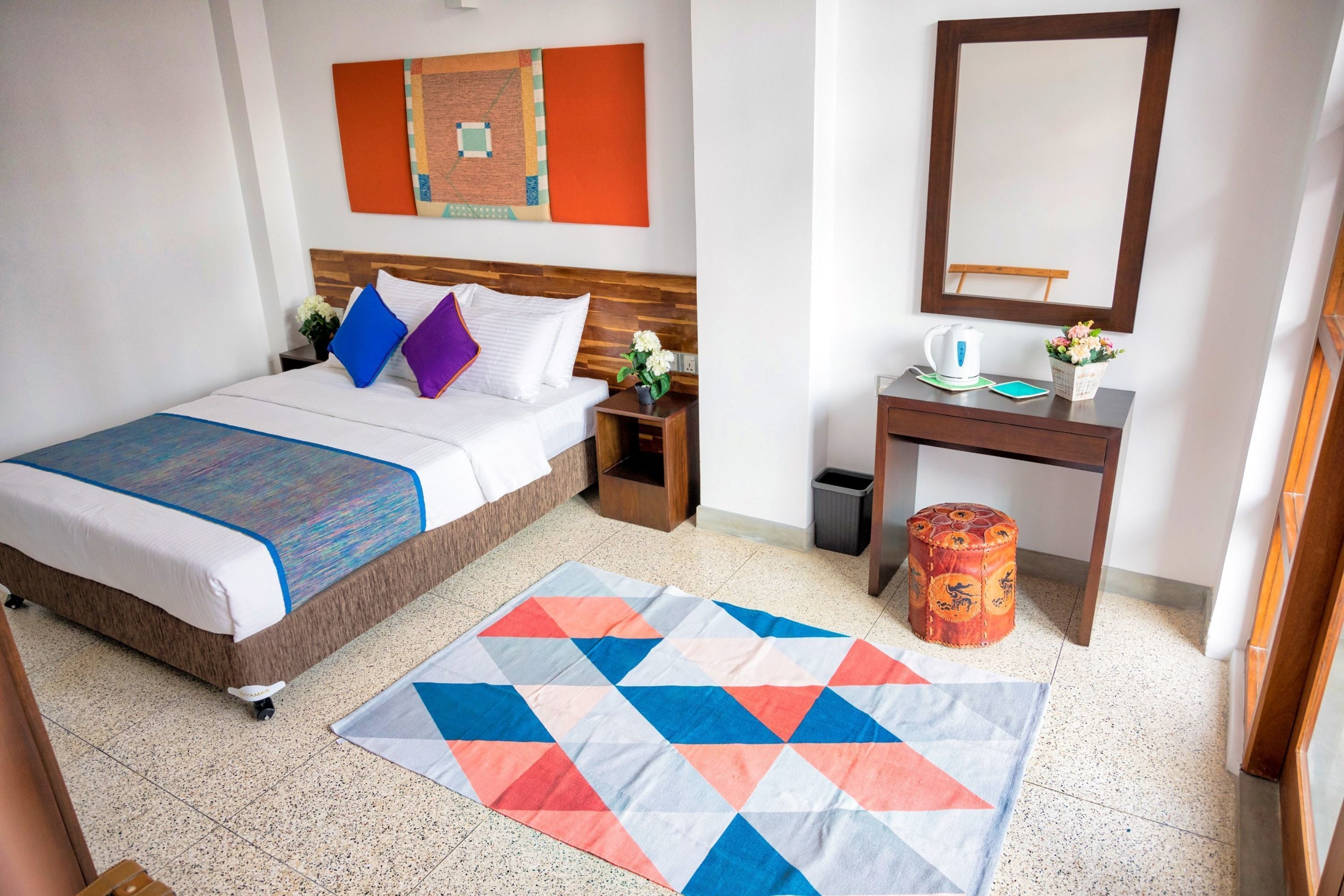 Habitación de hotel con cama, mesita de noche, pequeña mesa con espejo, cesto de basura y una alfombra colorida.