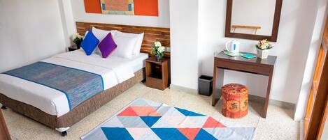 Habitación de hotel con cama, mesita de noche, pequeña mesa con espejo, cesto de basura y una alfombra colorida.