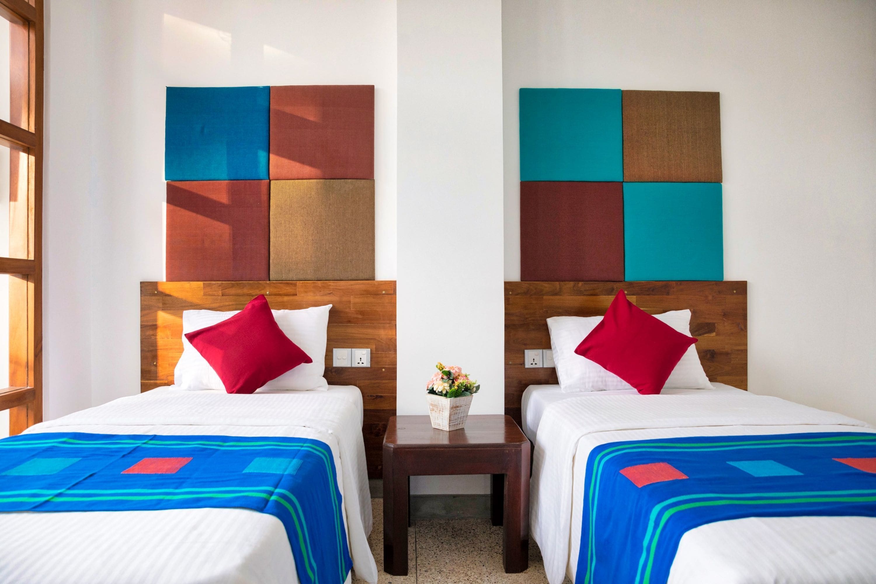 Habitación de hotel con dos camas, cada una con cabecero de madera, ropa de cama blanca y almohadas rojas. Una mesita con un florero se encuentra entre las camas.