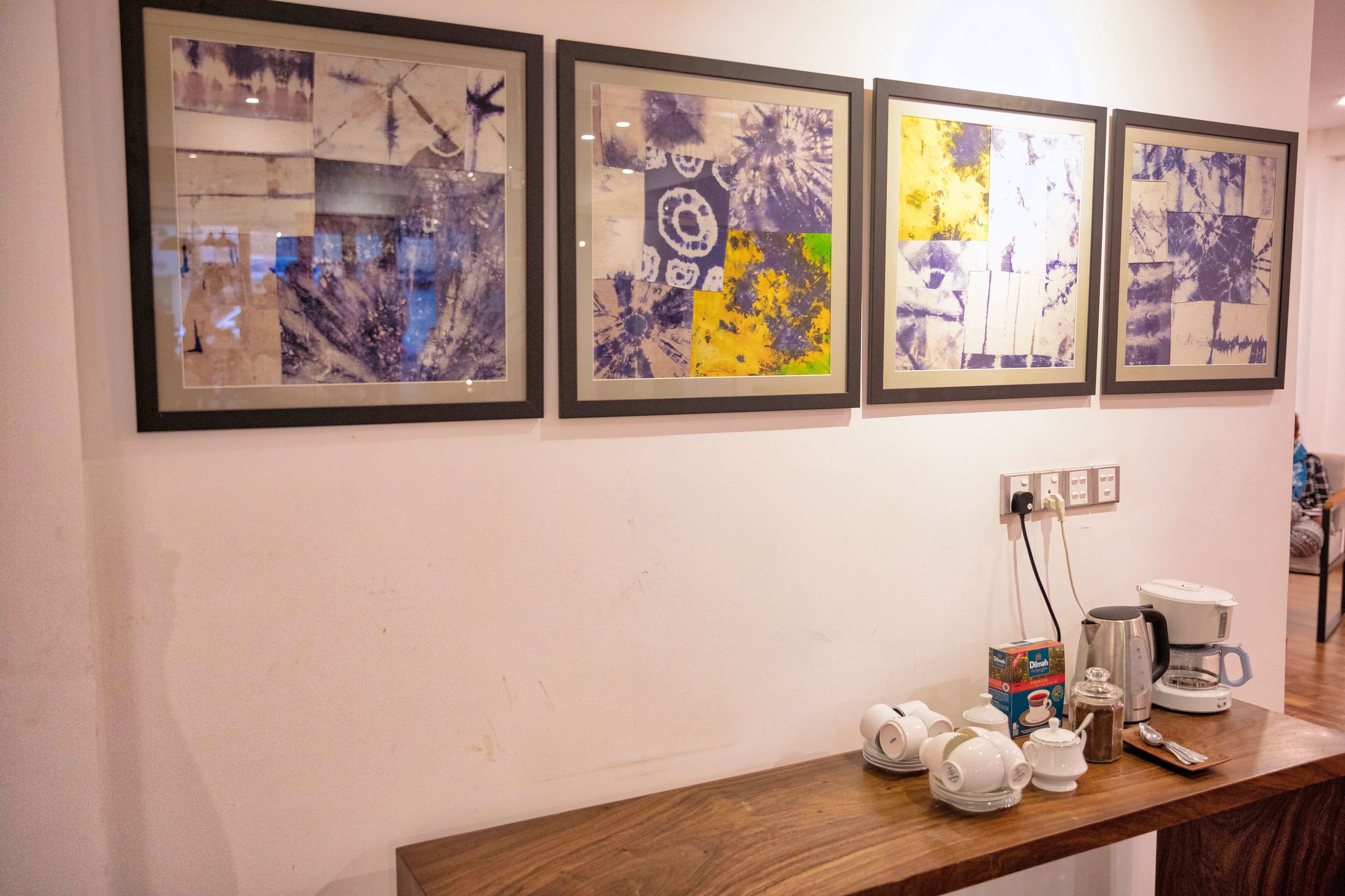 A wall with four framed abstract paintings and a wooden counter with a coffee maker, teapot, and cups.