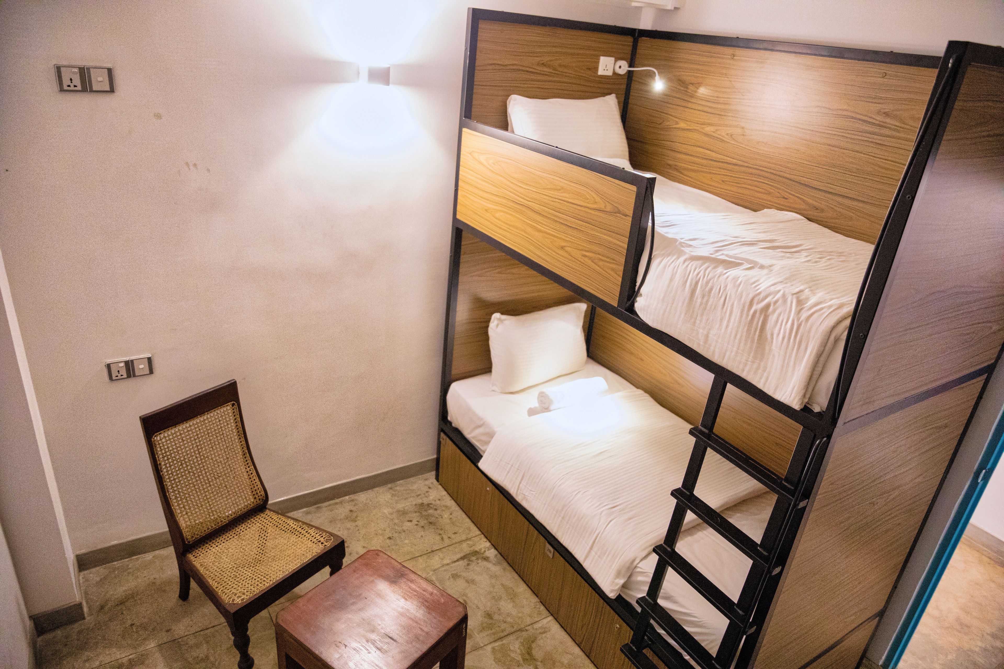 A bunk bed with a wooden headboard and white bedding, a wicker chair, and a small wooden table in a room with a plain wall and a light switch.