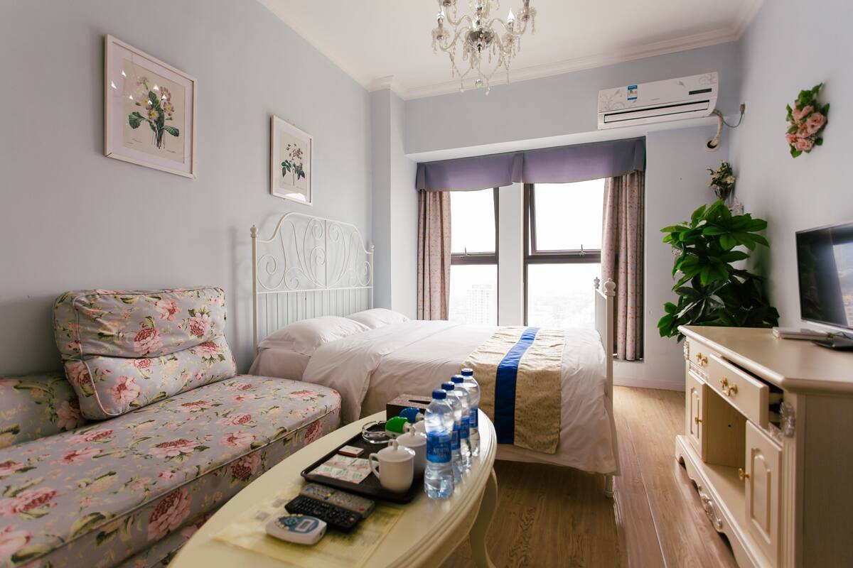 Habitación de hotel con cama, sofá, mesita de noche con bandeja de artículos de aseo, televisión y ventana con cortinas.