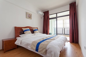 A bedroom with a wooden bed, white bedding, a blue and beige decorative strip, a nightstand, a wall-mounted air conditioner, and a large window with red curtains.