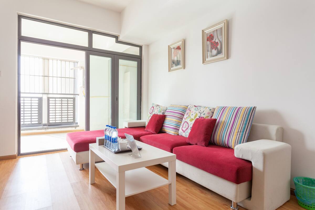 Un salón moderno con un sofá blanco, almohadas coloridas, una mesa de centro blanca y un puerta corrediza de vidrio que da a un balcón.