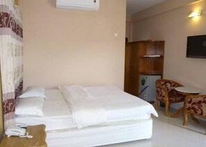 Quarto de hotel com uma cama, uma mesinha, uma cadeira e uma TV fixada na parede.