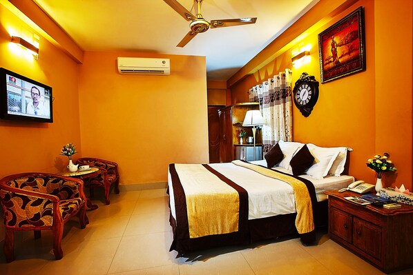 Quarto de hotel com cama, duas cadeiras, escrivaninha, relógio e televisão.