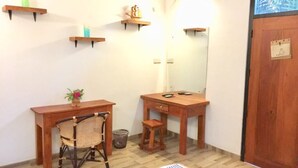 Une petite salle de bain compacte, avec un bureau et une chaise en bois, un petit tabouret et un miroir. On y trouve des étagères en bois au mur et une porte en bois.