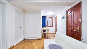 Um corredor com piso de madeira, paredes brancas, uma porta branca e outra de madeira, e um sofá branco com manta cinza.