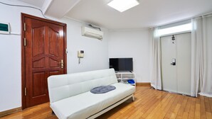 Quarto com piso de madeira, um sofá branco, uma TV sobre um móvel, um aparelho de ar condicionado e uma porta com cortina.