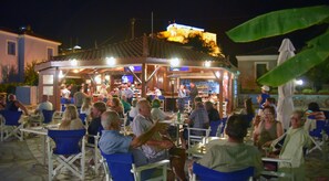 An outdoor bar area with tables and chairs, a stage for performances, and a building with a tiled roof in the background.