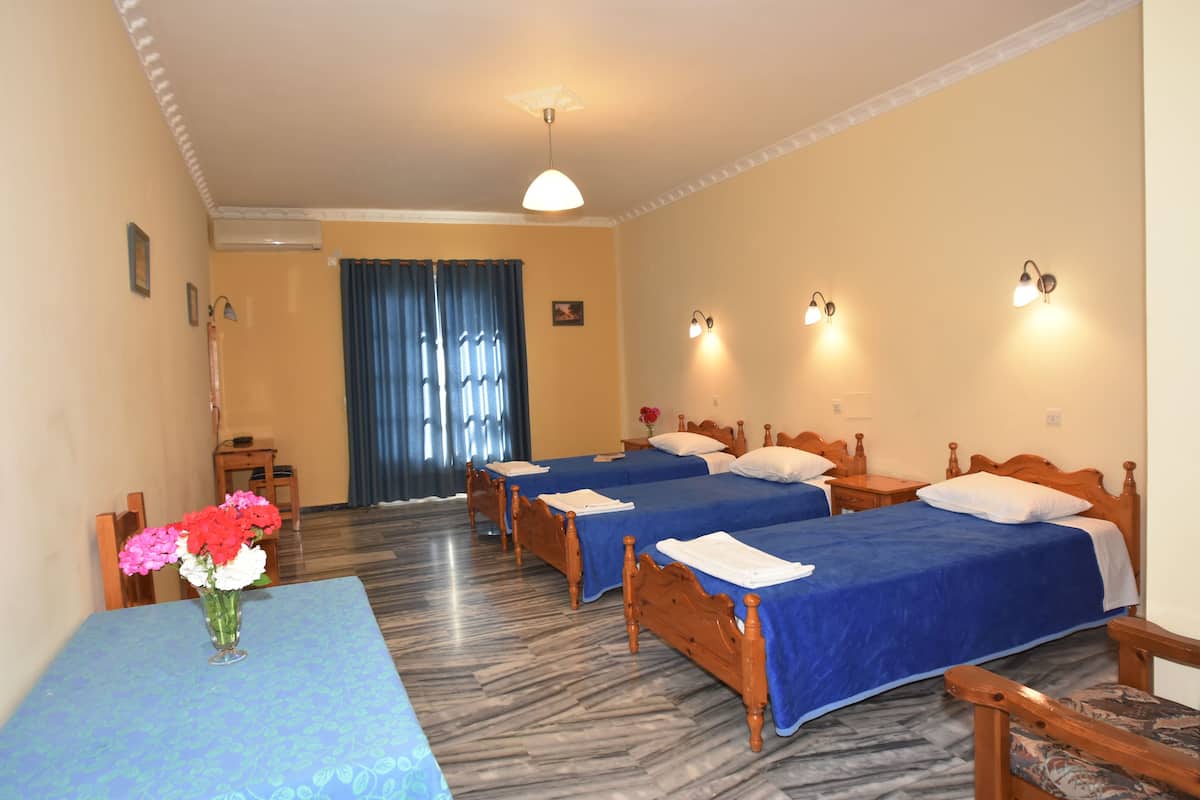 A hotel room with three wooden beds, a blue table with a vase of flowers, and a view of the outside through a window with blue curtains.