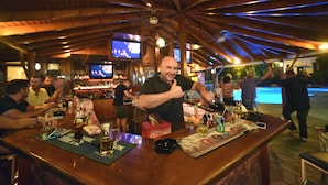 Zona bar con travi in legno, diverse televisioni e bancone con bicchieri e bottiglie.
