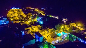 Vista aerea di una cittadina costiera di notte, con edifici illuminati, una collina in evidenza con una struttura simile a un forte e un'area piscina.
