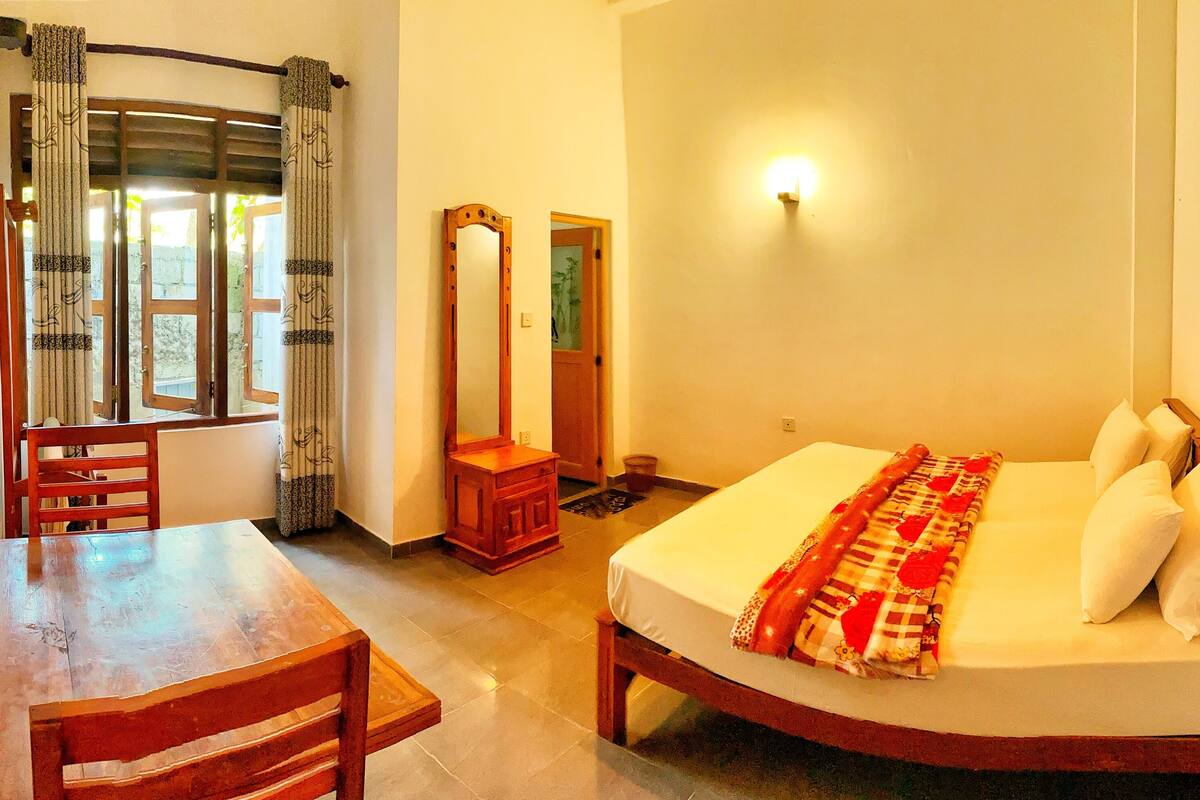 Une chambre à coucher avec un lit, une chaise en bois, une petite table, un miroir et une fenêtre avec des rideaux.