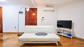 Ein Zimmer mit einem weißen Sofa, einem Fernseher auf einem weißen Schrank, einer Holztür und einer Klimaanlage an der Wand.