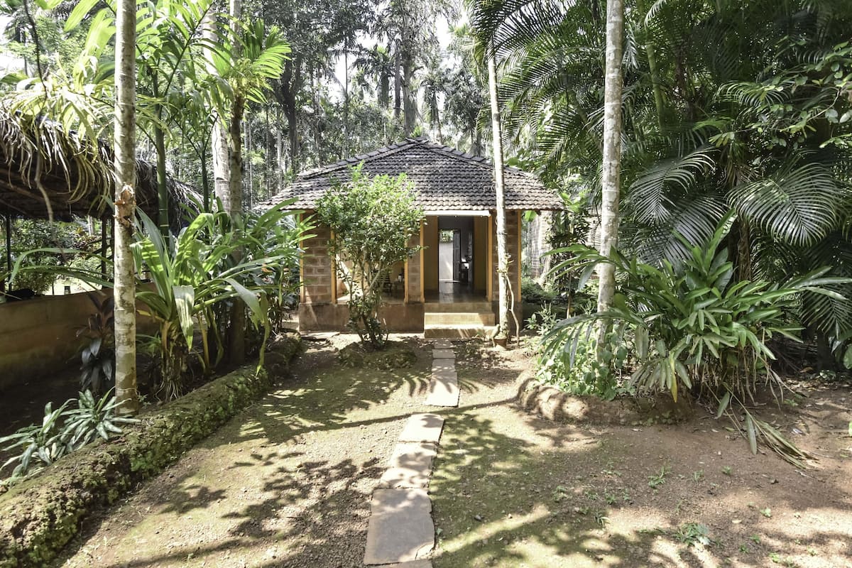 A small, rustic building surrounded by lush greenery, with a dirt path leading to the entrance.