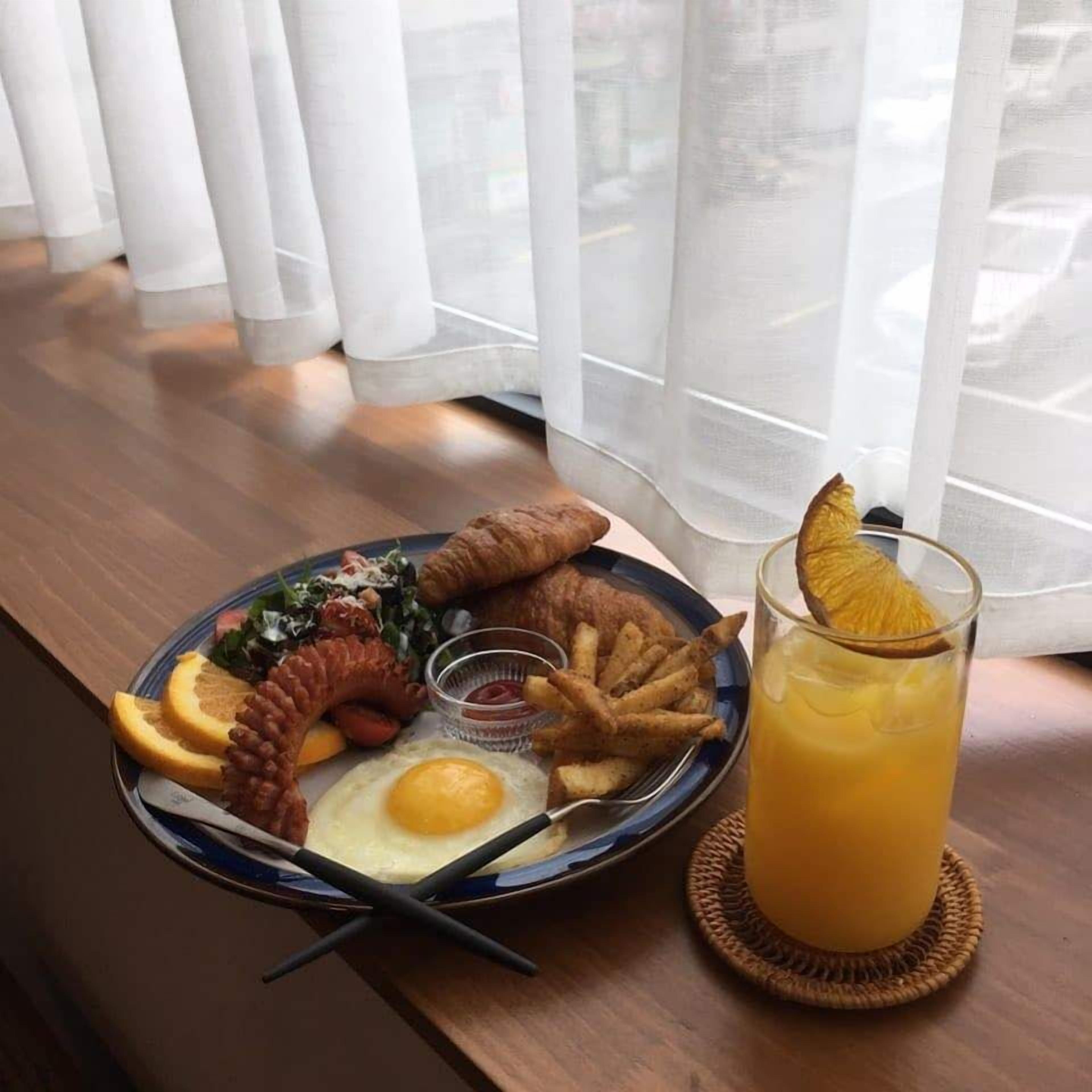 Une assiette de petit-déjeuner avec des œufs, des saucisses, des frites et un verre de jus d’orange, posée sur une table en bois près d’une fenêtre avec des rideaux.