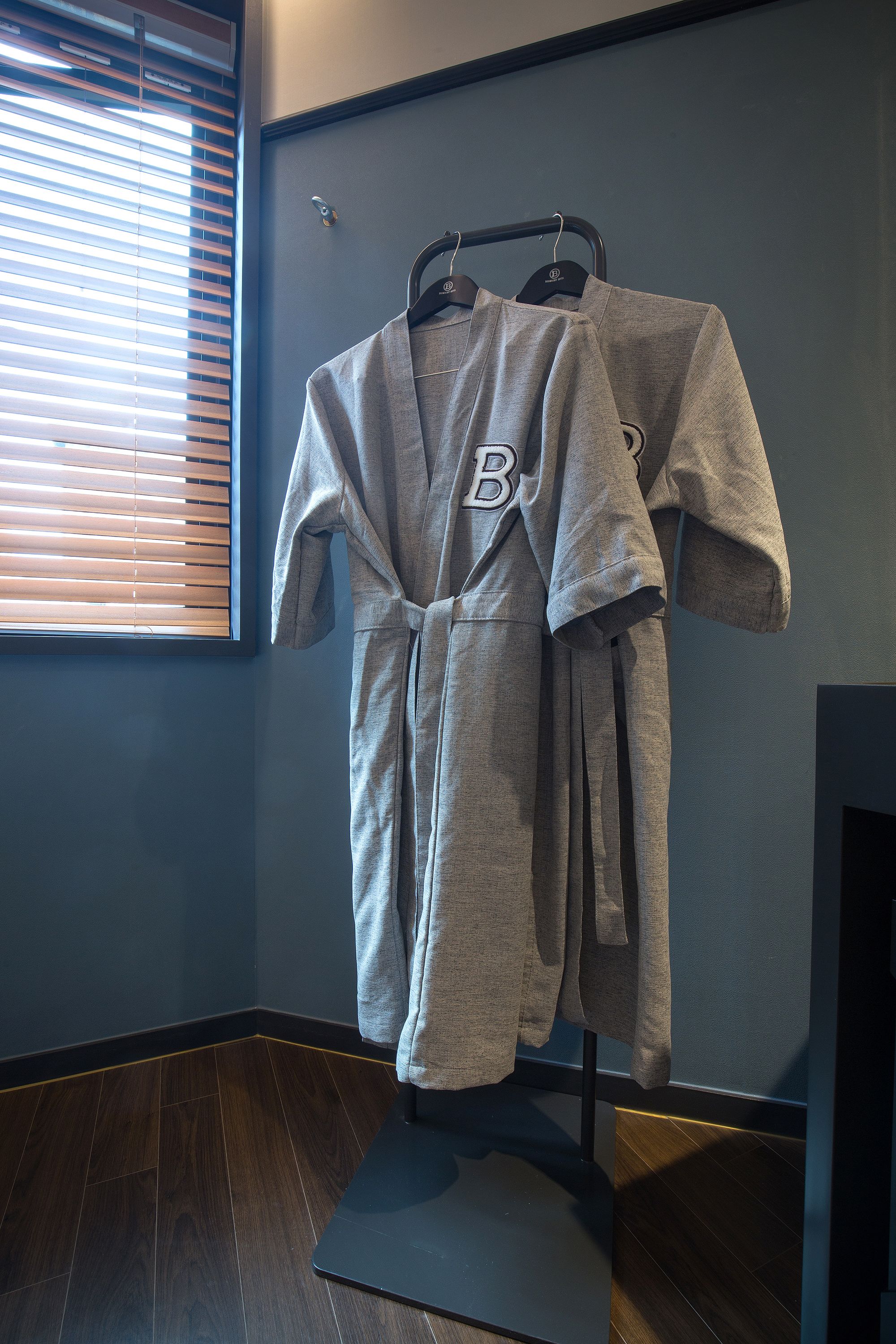 A bathroom with a wooden floor, a towel rack with two bathrobes, and a window with blinds.