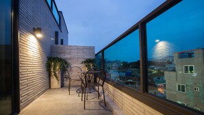 Un balcon offrant une vue sur la ville au crépuscule, avec une table et des chaises, un pot de fleurs et une balustrade en verre.