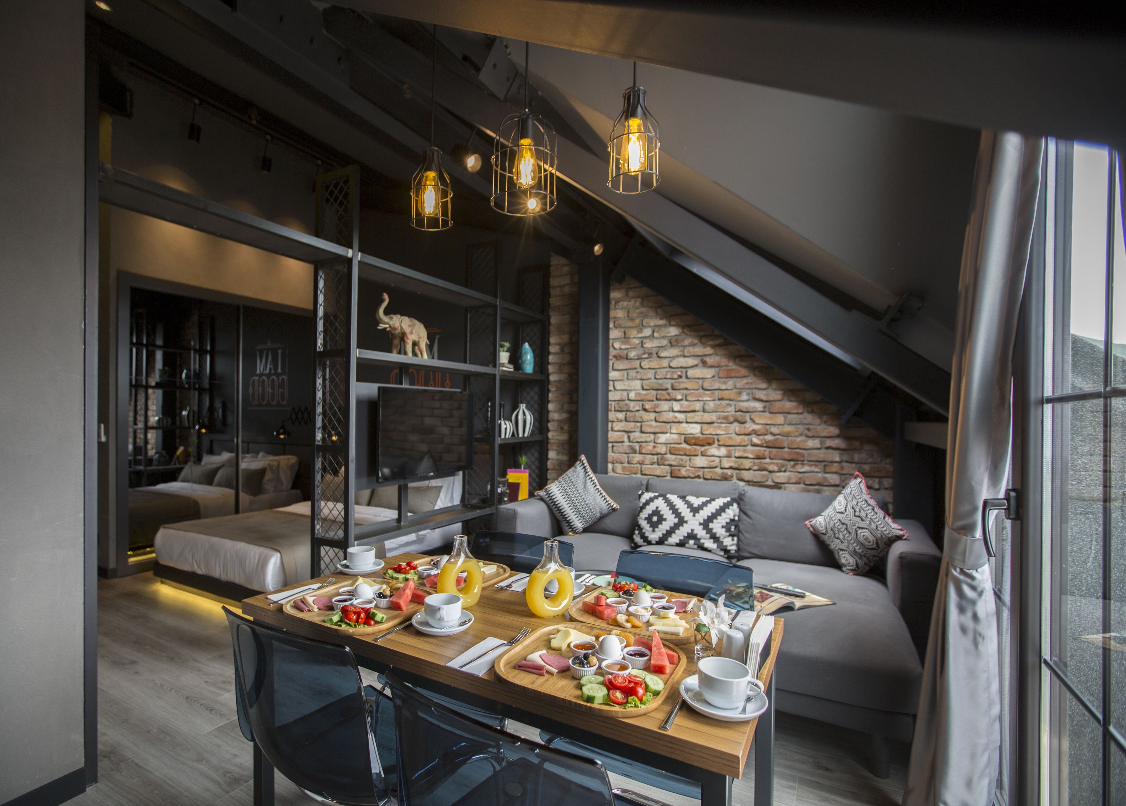 Un comedor moderno con una mesa de madera dispuesta para una comida, un sofá con cojines estampados y una pared de ladrillo al fondo.