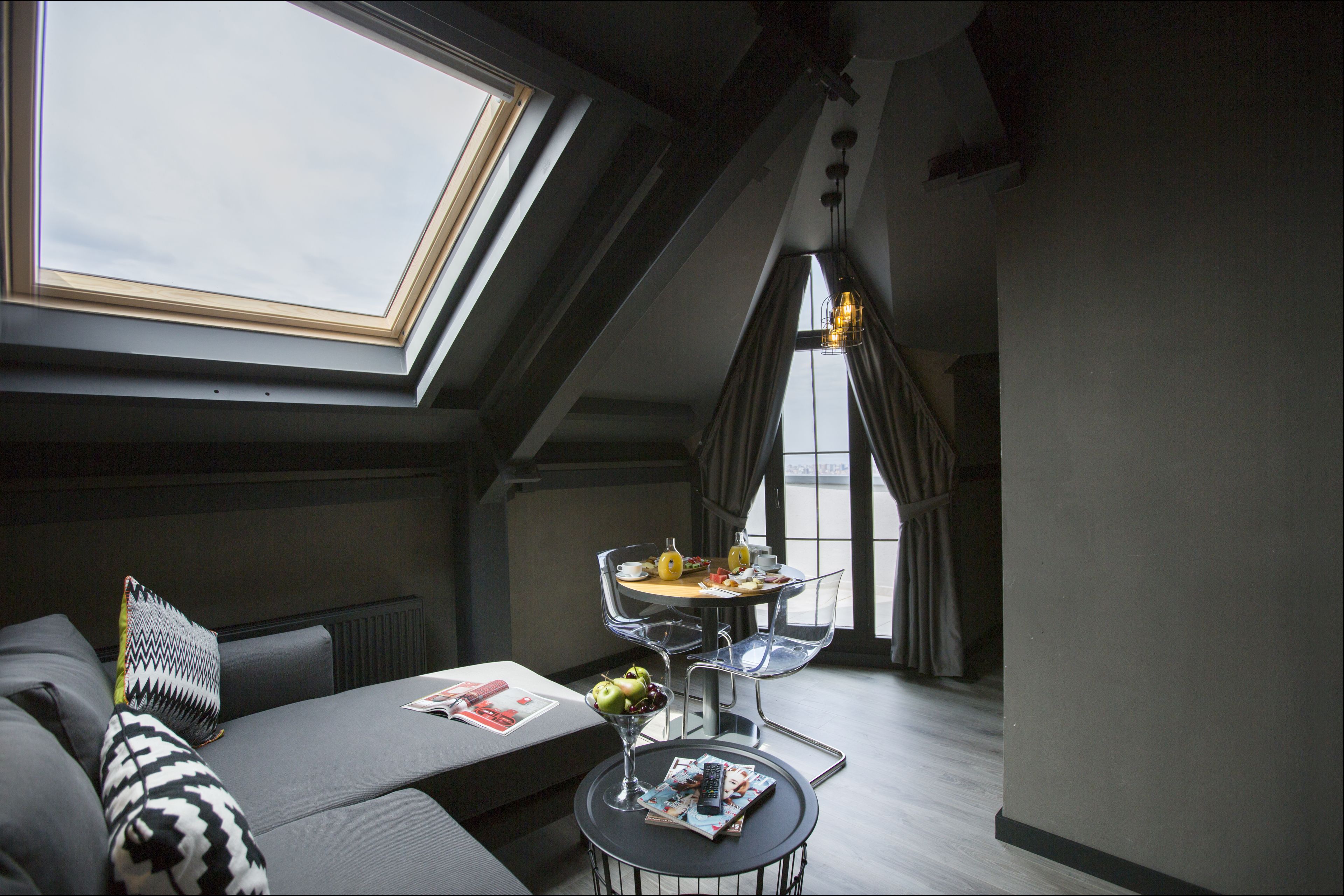 A modern loft bedroom with a skylight, a dining area with a table and chairs, and a cozy seating area with a sofa and pillows.
