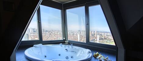 A modern bathroom with a large bathtub, a window offering a city view, and a towel on the counter.