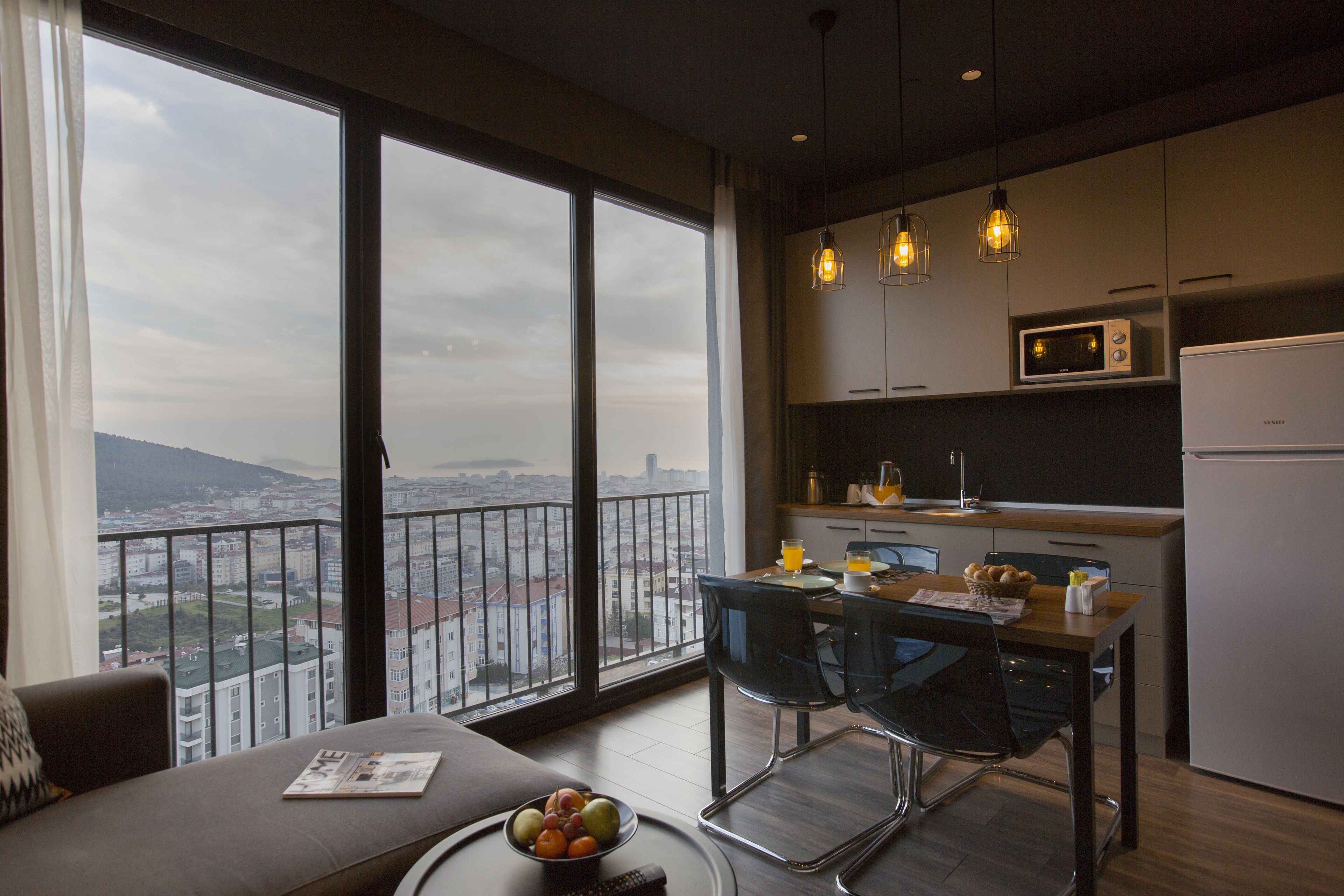 Una cocina moderna con vistas a un paisaje urbano, una zona de comedor con mesa y sillas, y un sofá con una revista.