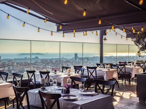 Un área de comedor al aire libre con mesas preparadas para dos personas, con vistas a un paisaje urbano.