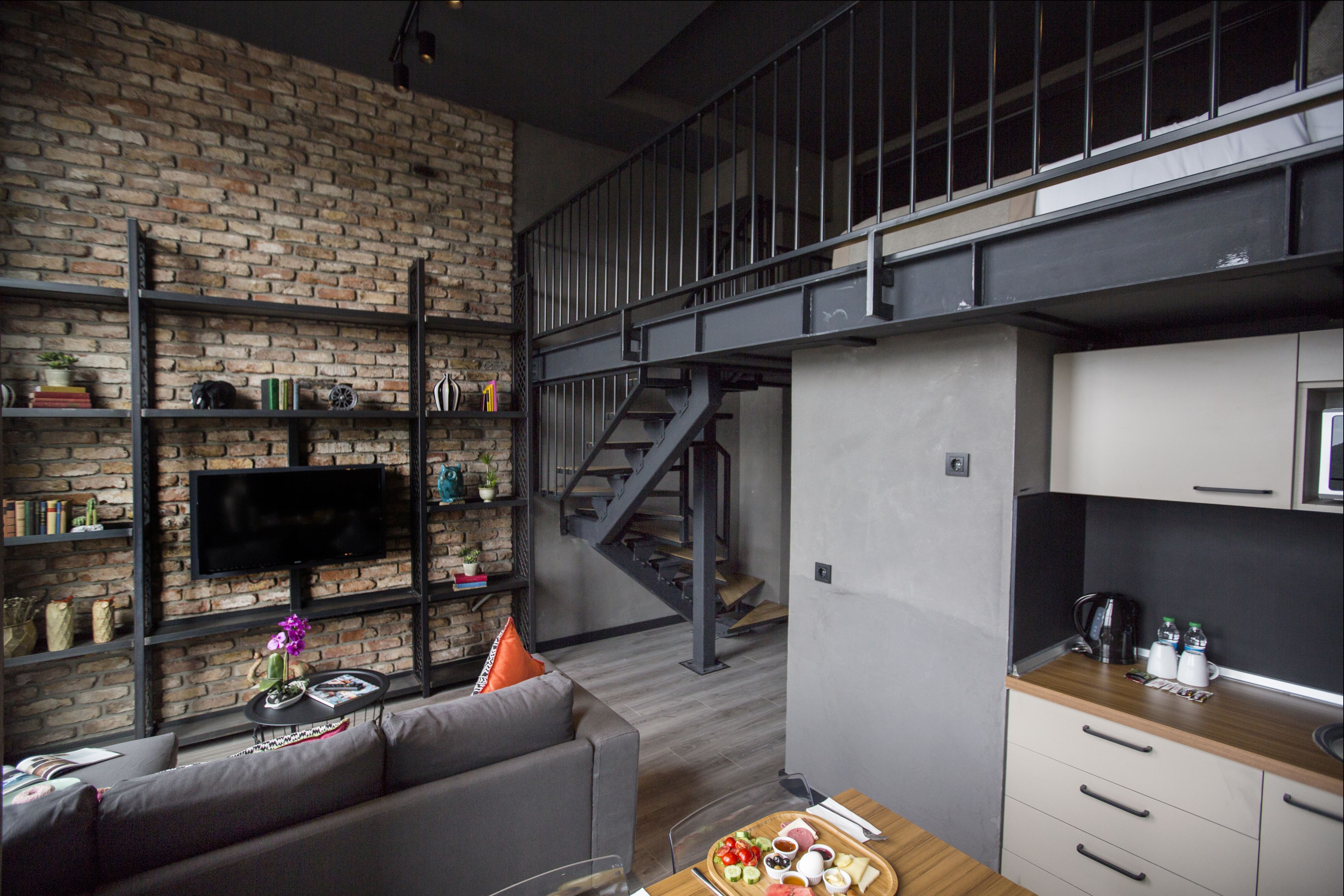 A modern living space with a brick wall, a flat-screen TV, a sofa, a dining table with a tray of food, and a staircase leading to an upper level.