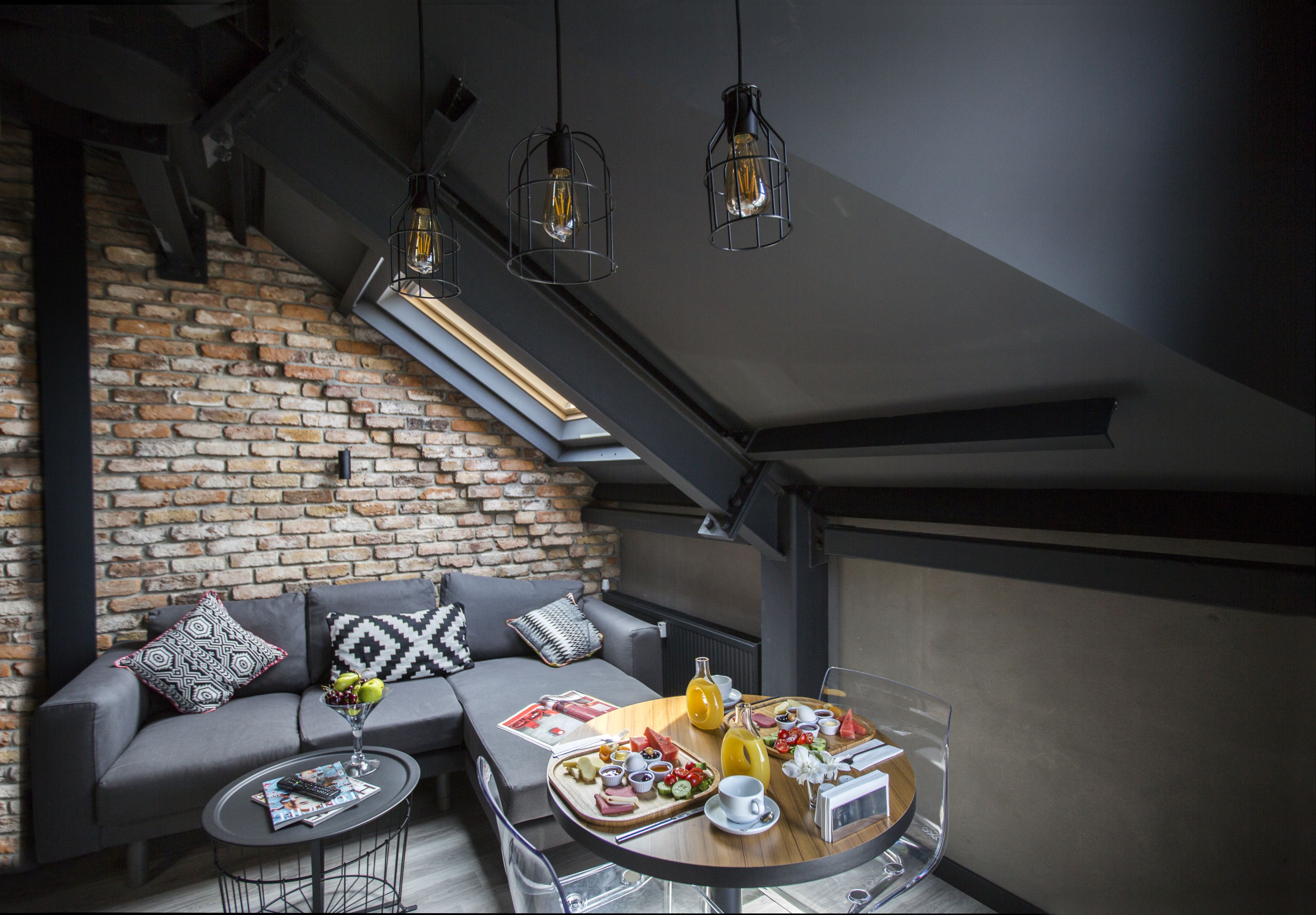 Un intérieur moderne avec un mur en briques, un canapé gris agrémenté de coussins à motifs, une petite table ronde sur laquelle reposent un magazine et une télécommande, et un plateau de petit-déjeuner posé sur une table en verre.