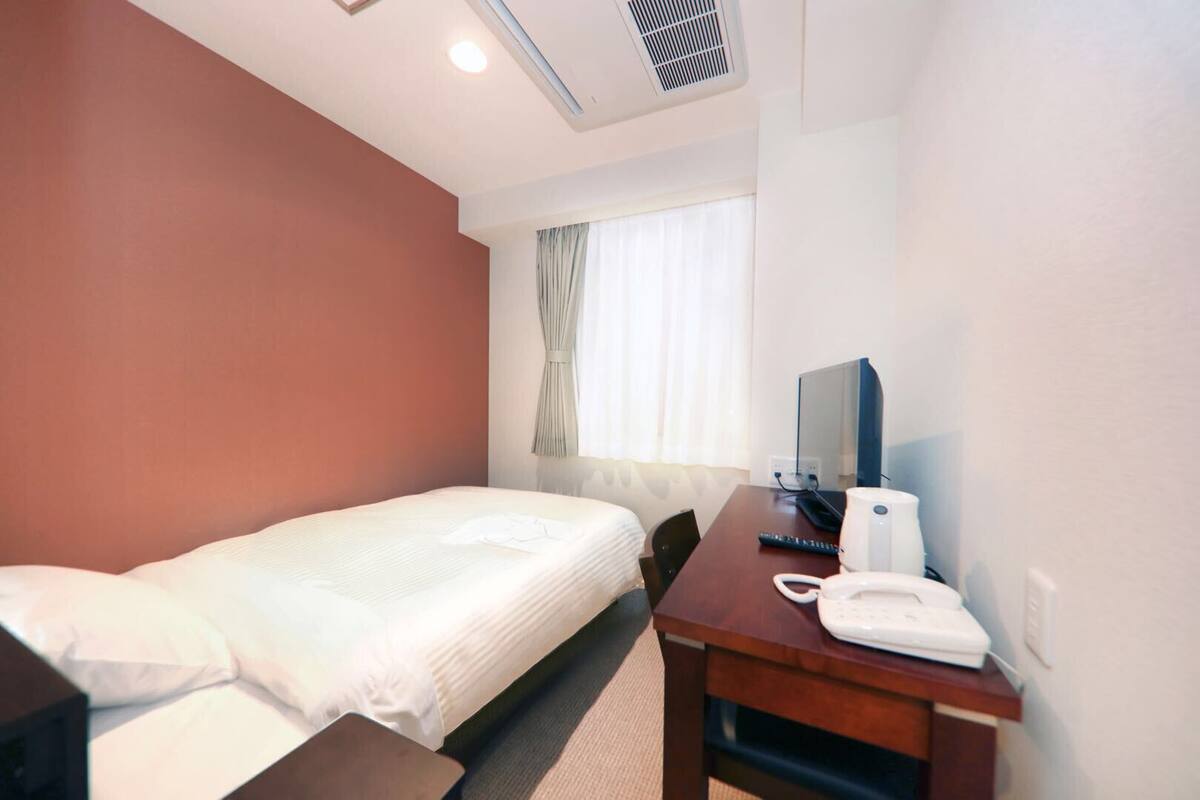 A hotel room with a single bed, a desk with a telephone and a computer, and a window with curtains.