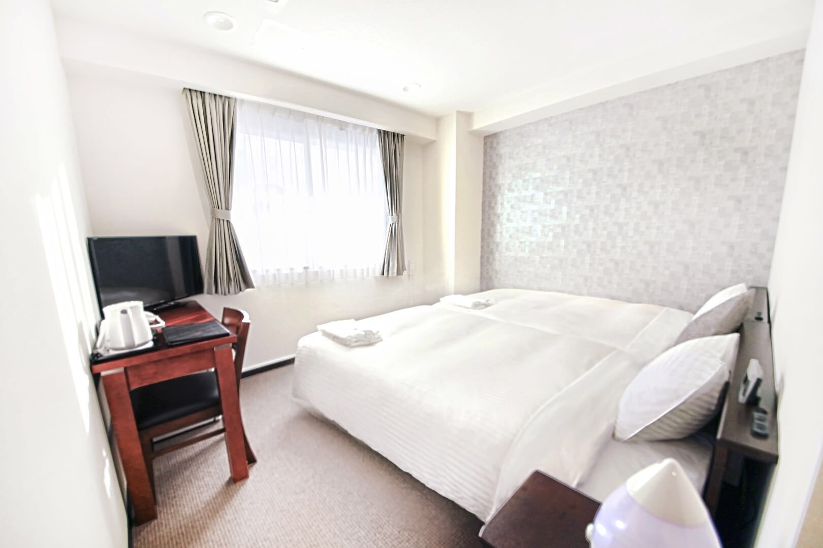 A hotel room with a bed, a TV, a desk with a coffee maker, and a window with curtains.
