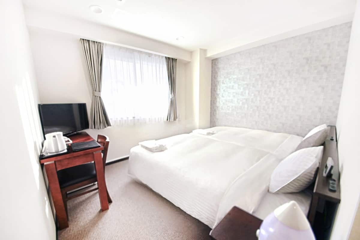 Habitación de hotel con cama, televisión, escritorio con cafetera y ventana con cortinas.