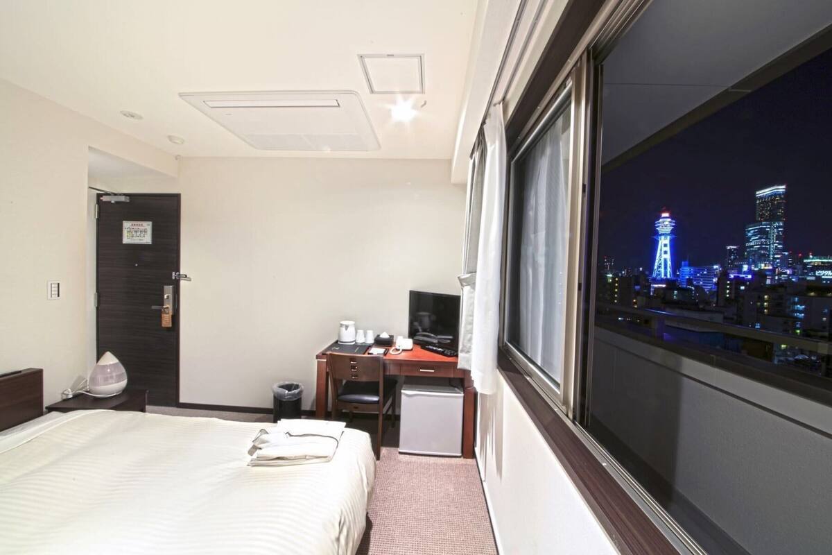 A hotel room with a bed, desk, and a view of a cityscape at night.