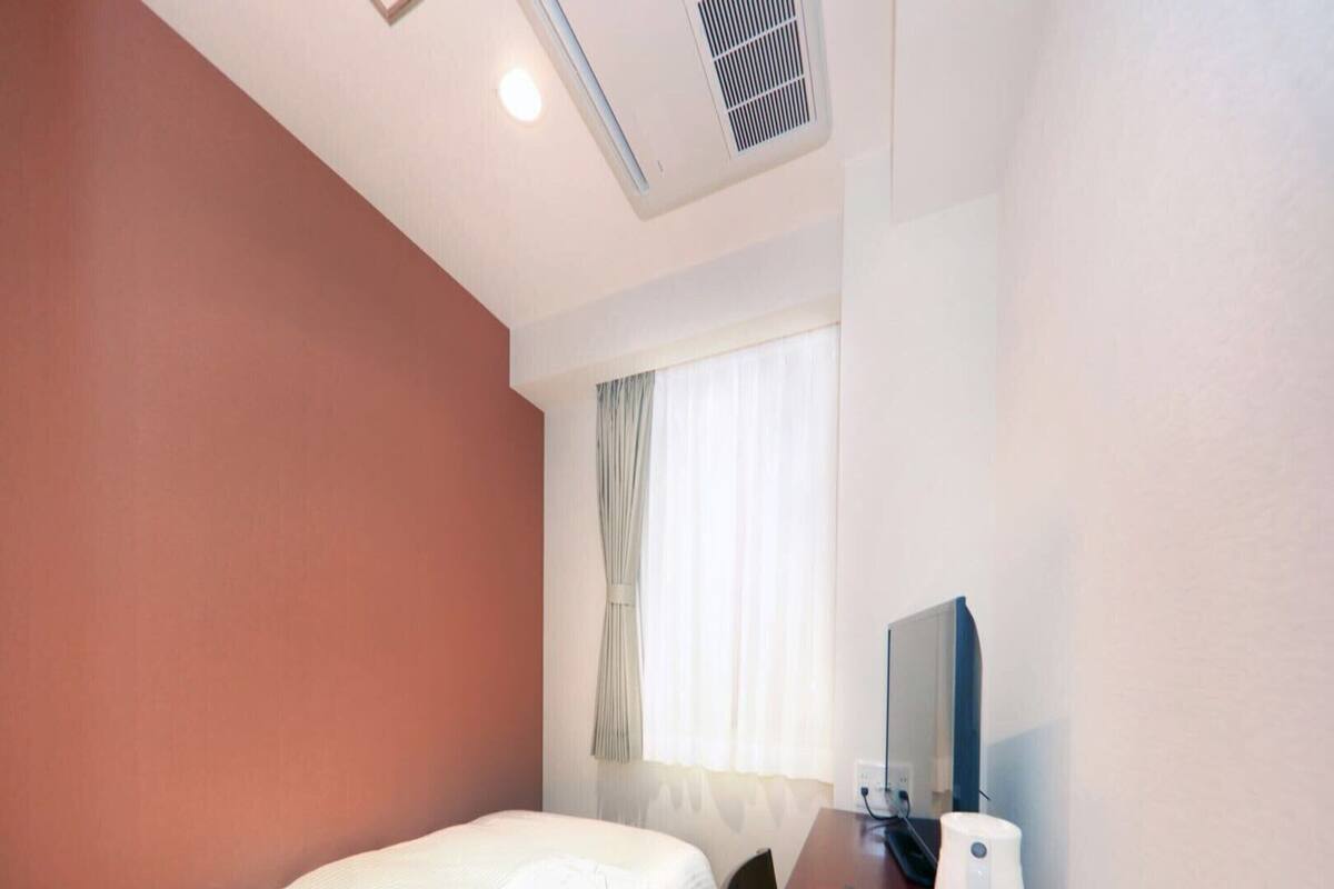 A hotel room with a single bed, a desk with a telephone and a remote control, a mirror, and a window with curtains.